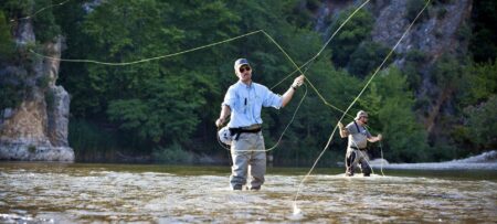 The image shows a man fly fishing in a river wearing waders and casting his line.