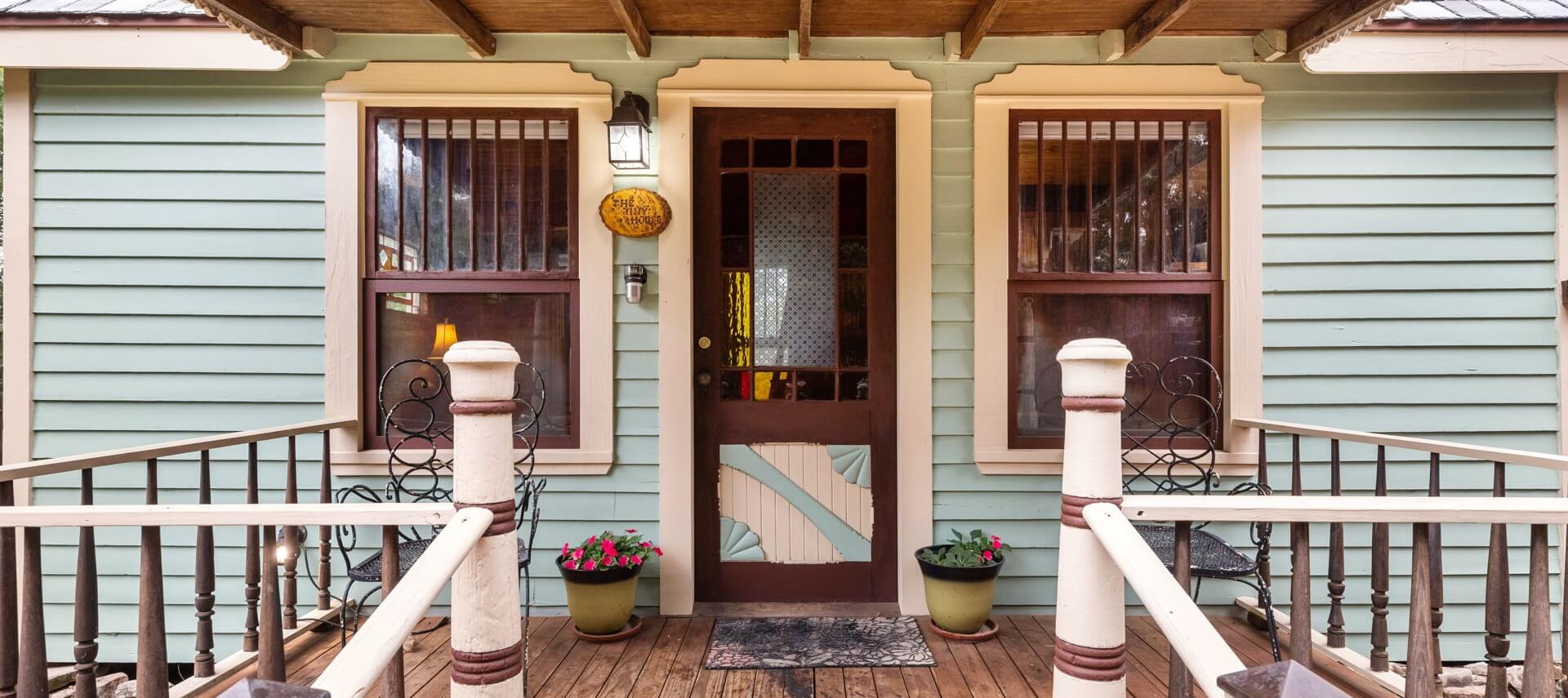 Close up of front porch of a tiny house with black chairs, a colorful front door and two windows.