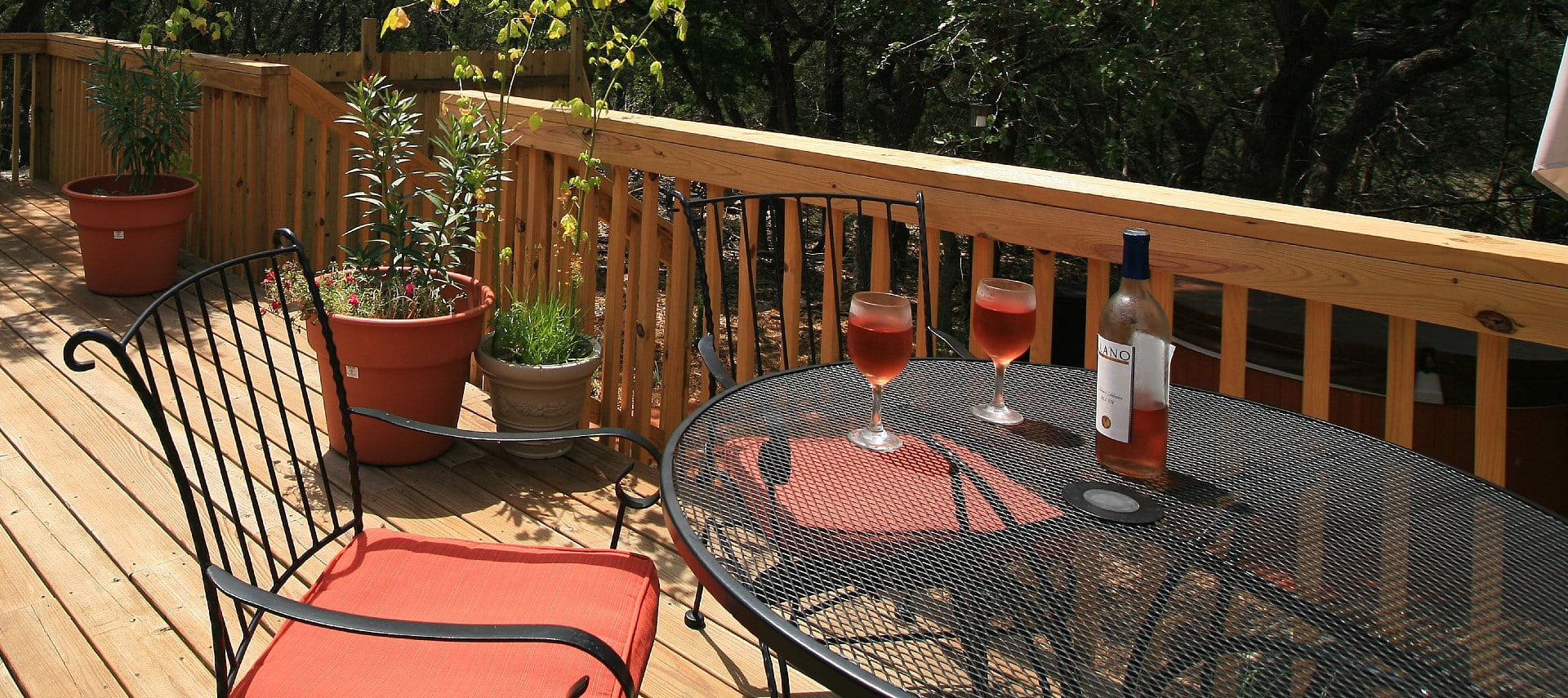 A wrought iron patio table with two chairs, bottle of wine and two glasses near potted plants on a deck