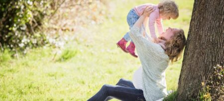 A mothers sitting against a tree lifting up her child in a park.
