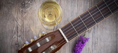 An image showing a glass of white wine next to a guitar and purple flower.