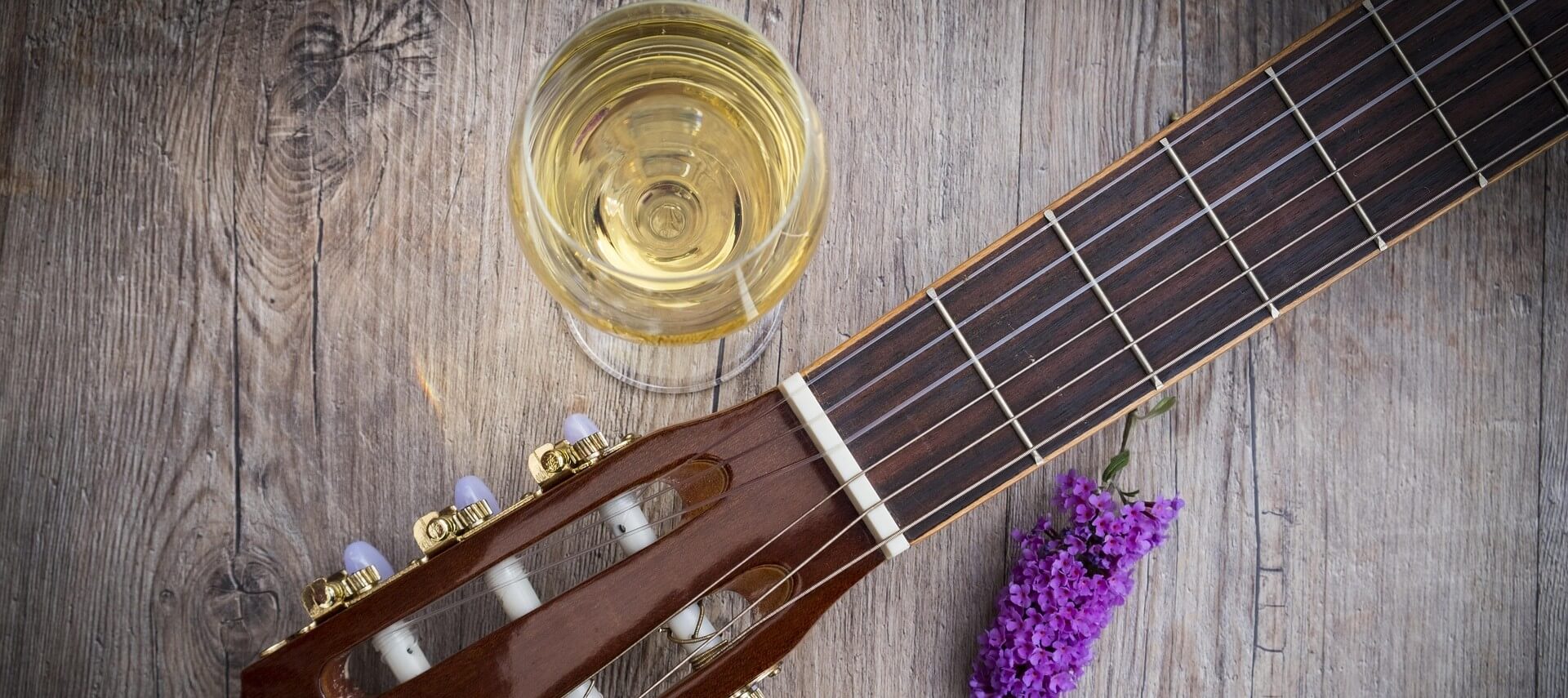 An image showing a glass of white wine next to a guitar and purple flower.